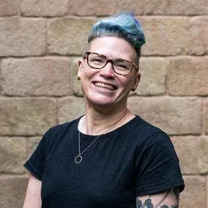 A woman wearing glasses, short cropped blue hair, a black tshirt and necklace smiles in the camera for a headshot. The background is a red brick wall.