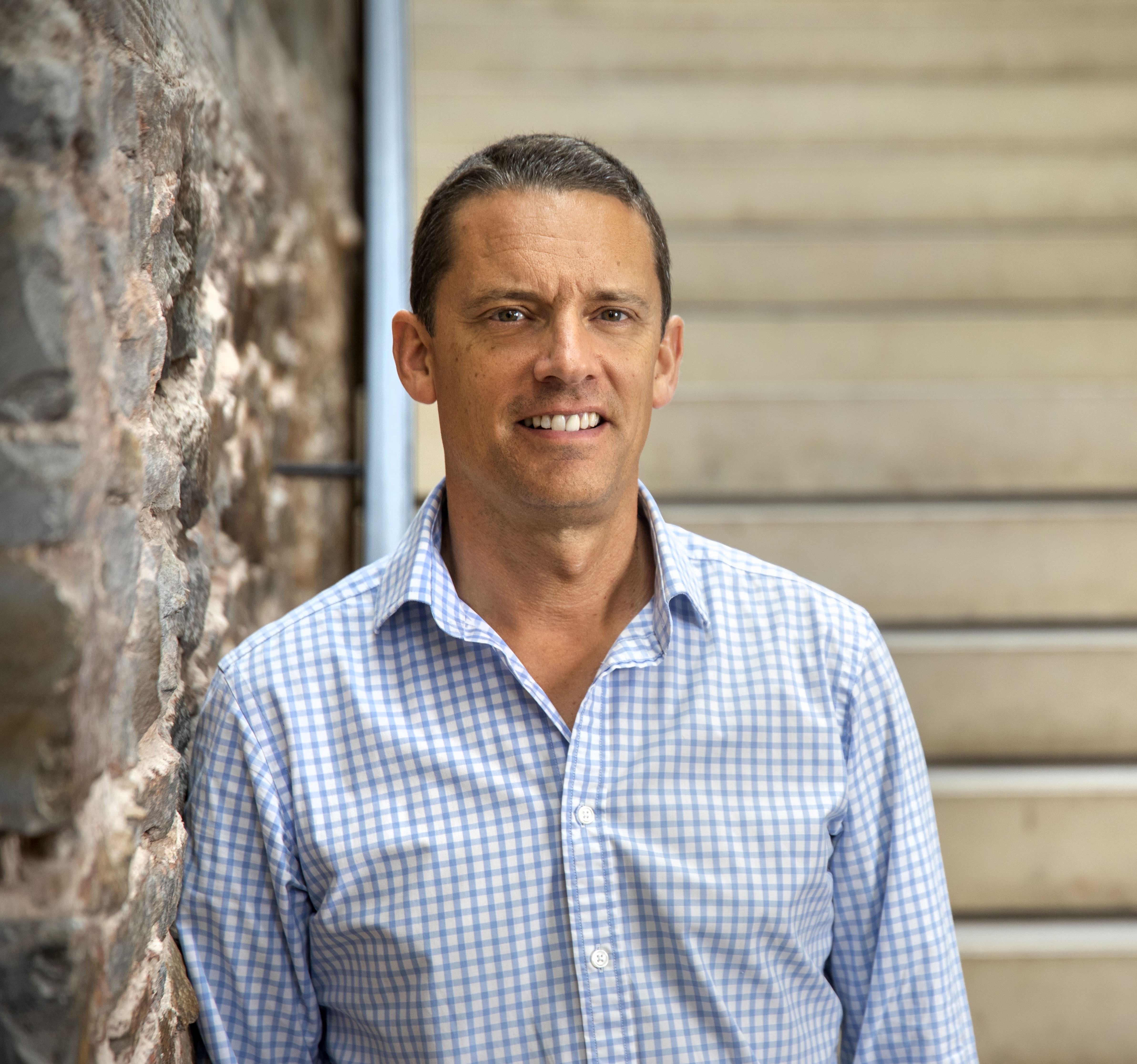 A headshot of a man, Bevis Watts, with short brown hair wearing a blue and white checked shirt facing the camera